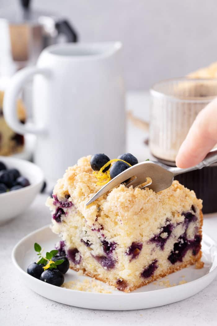 Fork about to take a bite from a slice of blueberry coffee cake on a white plate.