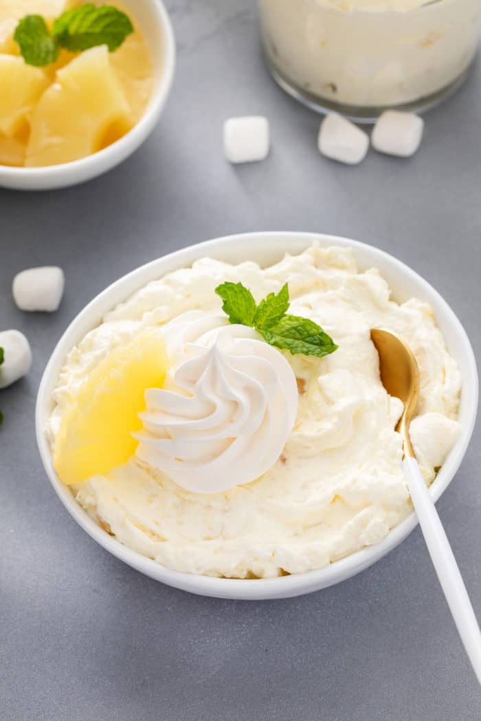 Spoon in a white bowl of pineapple fluff garnished with whipped cream and a piece of pineapple.