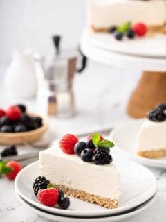 Slice of no-bake frozen cheesecake plated on a white plate, topped with mixed fresh berries. Additional slices of cheesecake are in the background