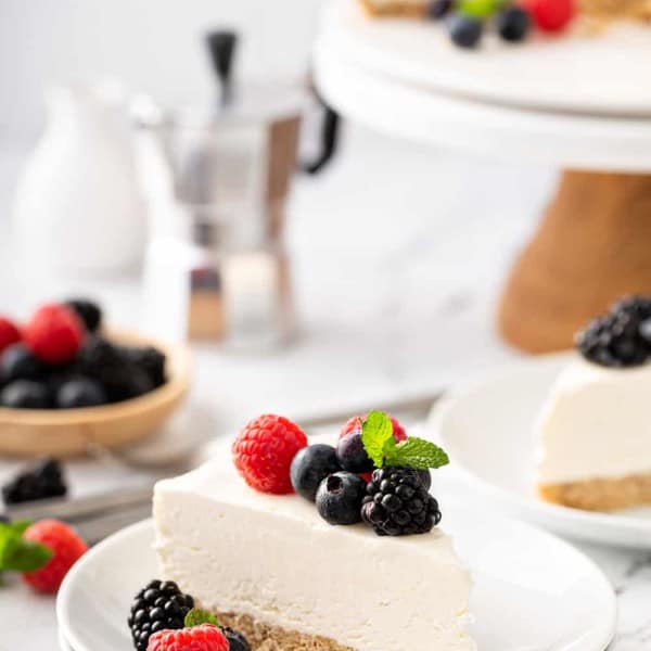 Slice of no-bake frozen cheesecake plated on a white plate, topped with mixed fresh berries. Additional slices of cheesecake are in the background
