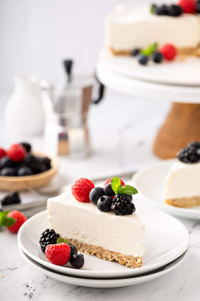 Slice of no-bake frozen cheesecake plated on a white plate, topped with mixed fresh berries. Additional slices of cheesecake are in the background