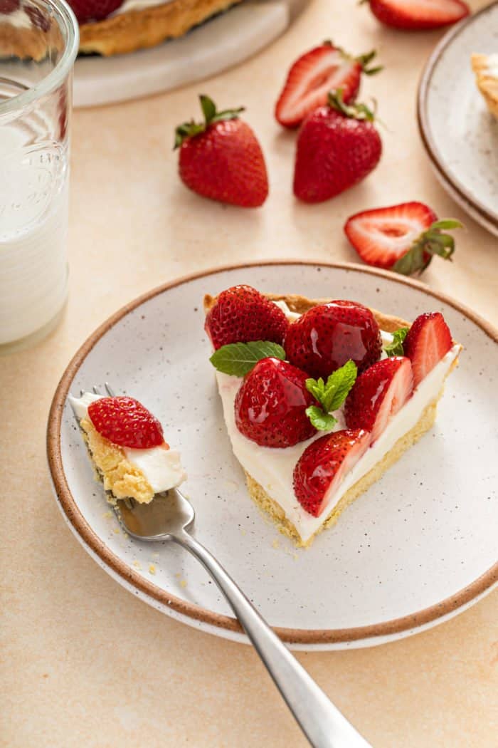 Fork taking a bite from a piece of strawberry tart on a white plate.