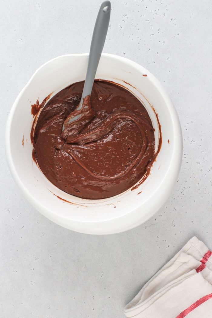 Chocolate zucchini bread batter being stirred with a spatula in a white mixing bowl.