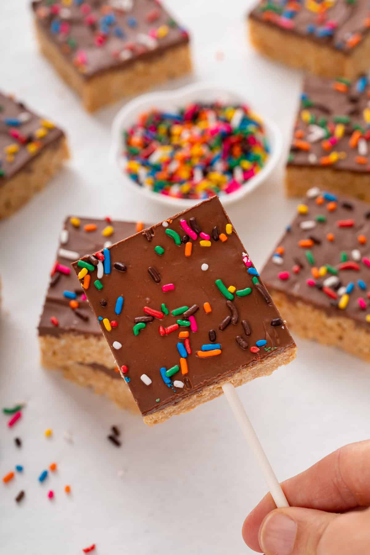 Hand holding up a sprinkle-topped scotcheroo bar on a lollipop stick.