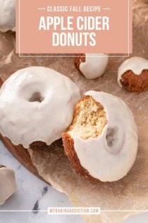 Two apple cider donuts on a piece of parchment paper. A bite has been taken from one of them. Text overlay includes recipe name.