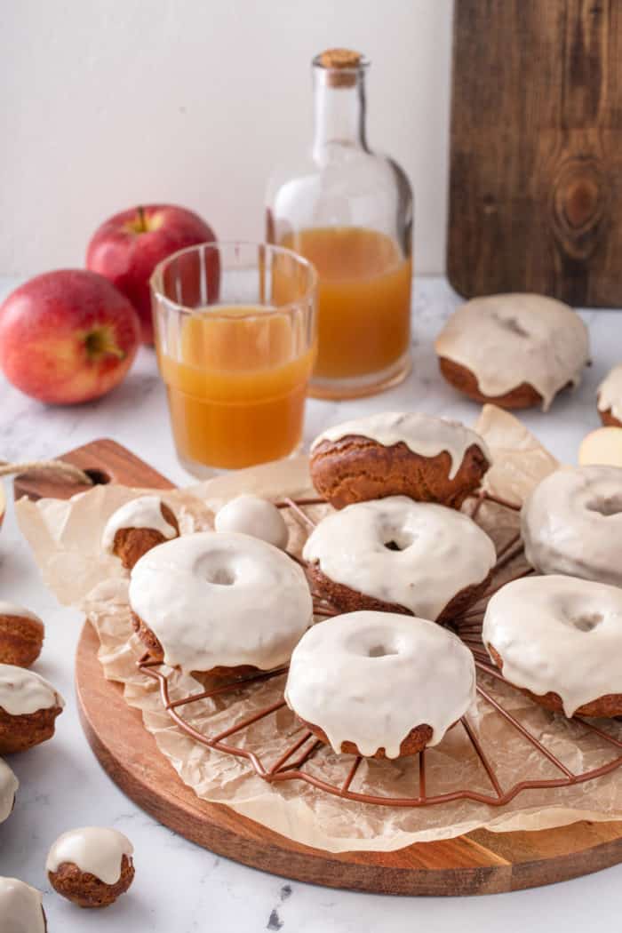 Glazed apple cider donuts cooling on a wire rack. A glass of cider and whole apples are in the background.