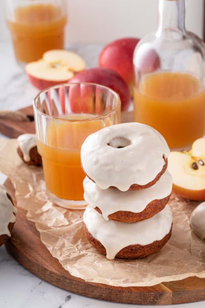 Three apple cider donuts stacked on a piece of parchment paper next to a glass of apple cider.