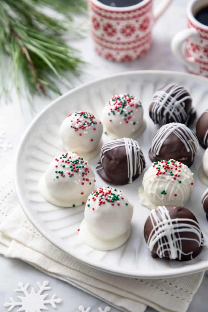 Close up of a white plate filled with white and dark chocolate-covered peanut butter balls. Red and white mugs of coffee are in the background.