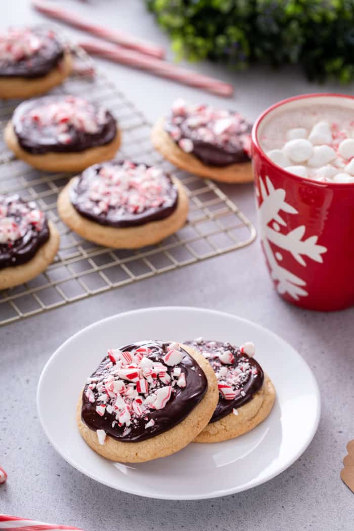 Two ganache-topped peppermint cookies on a white plate, with a wire rack of cookies in the background.