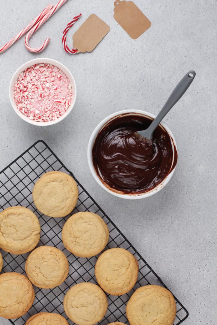 Baked peppermint cookies cooling on a wire rack, set next to a bowl of chocolate ganache and a bowl of crushed candy canes.