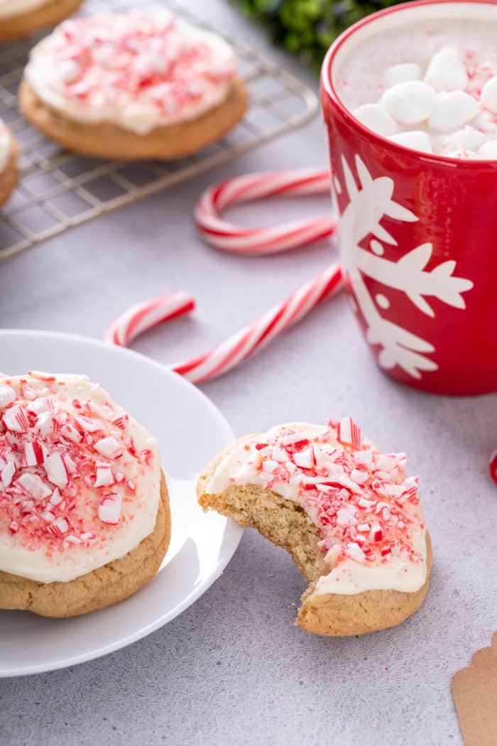 Cream cheese frosted peppermint cookie with a bite taken out of it leaning against a white plate holding a second peppermint cookie.