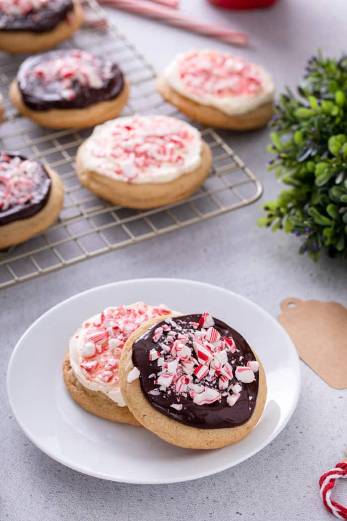 Cream cheese frosted peppermint cookie and ganache topped peppermint cookie, both decorated with chopped candy canes, on a white plate with more cookies in the background.