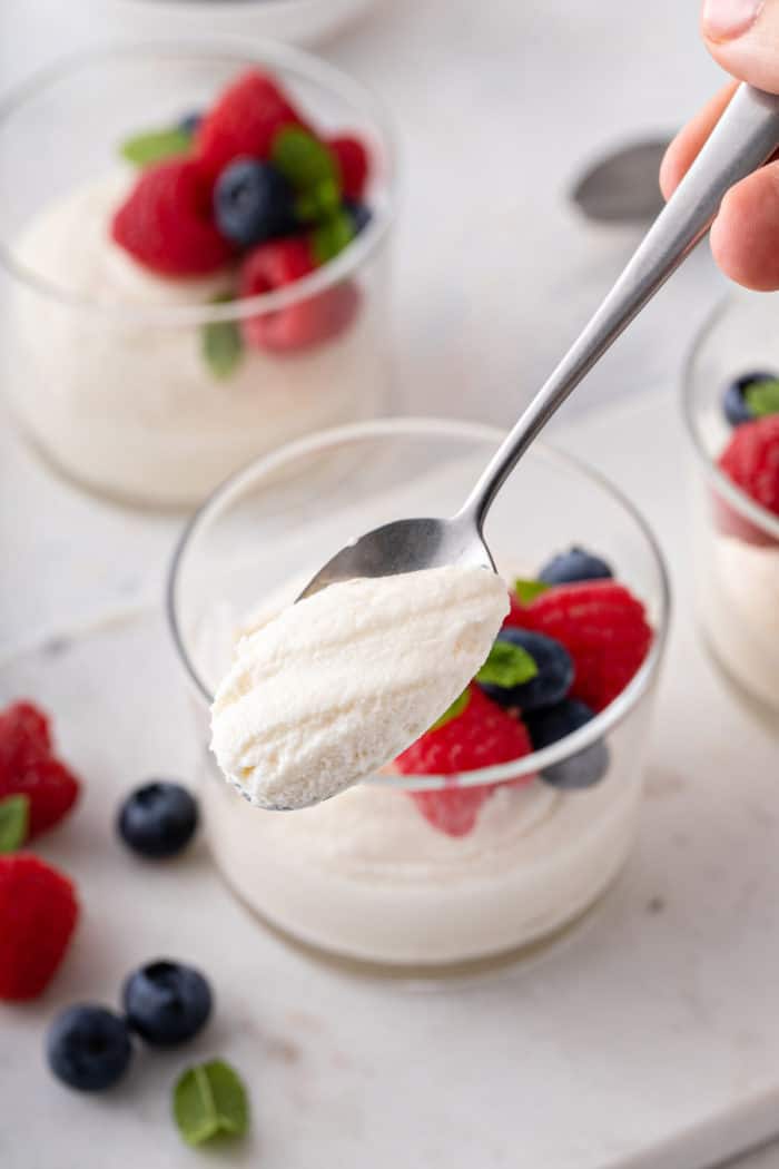 Spoon holding up a bite of white chocolate mousse. Glass dishes of the mousse are visible in the background.