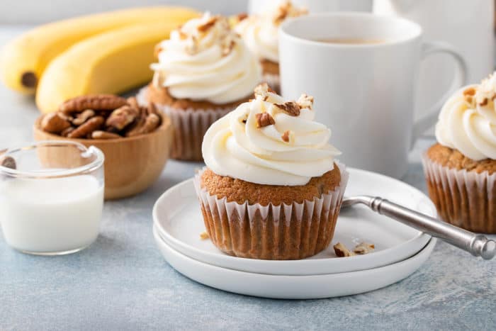 Plated banana cupcake topped with cream cheese frosting and chopped pecans. A cup of coffee and bananas are in the background