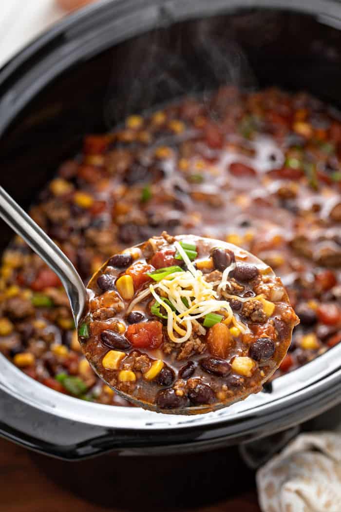 Ladle holding a spoonful of taco soup topped with shredded cheese, with the crock full of soup in the background