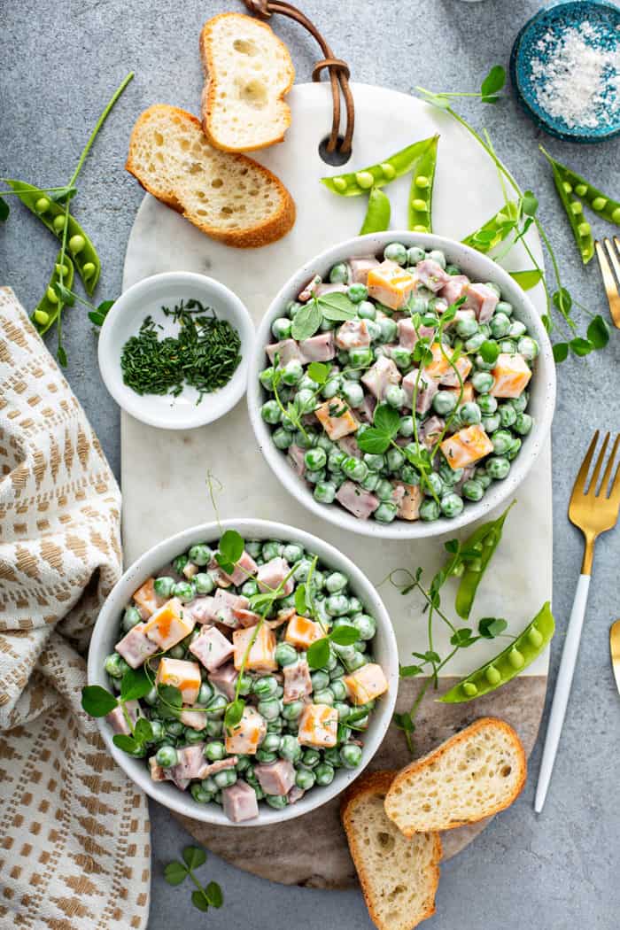 Overhead view of two small bowls of Amish pea salad on a white board surrounded by fresh peas, chives, and baguette slices