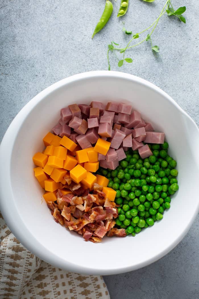 Green peas, cheddar cheese, ham, and bacon in a white mixing bowl