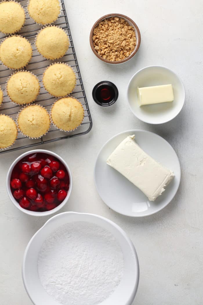 Baked cupcakes on a cooling rack next to the topping ingredients for cherry cheesecake cupcakes.