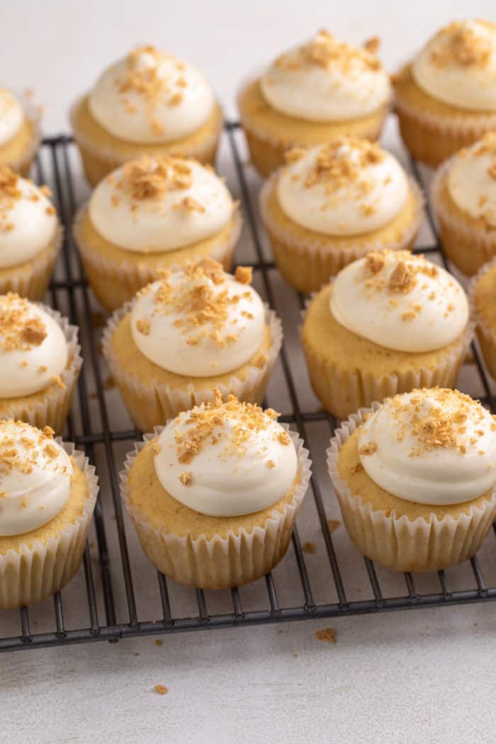 Graham cracker cupcakes topped with cream cheese frosting and graham cracker crumbs on a wire cooling rack.
