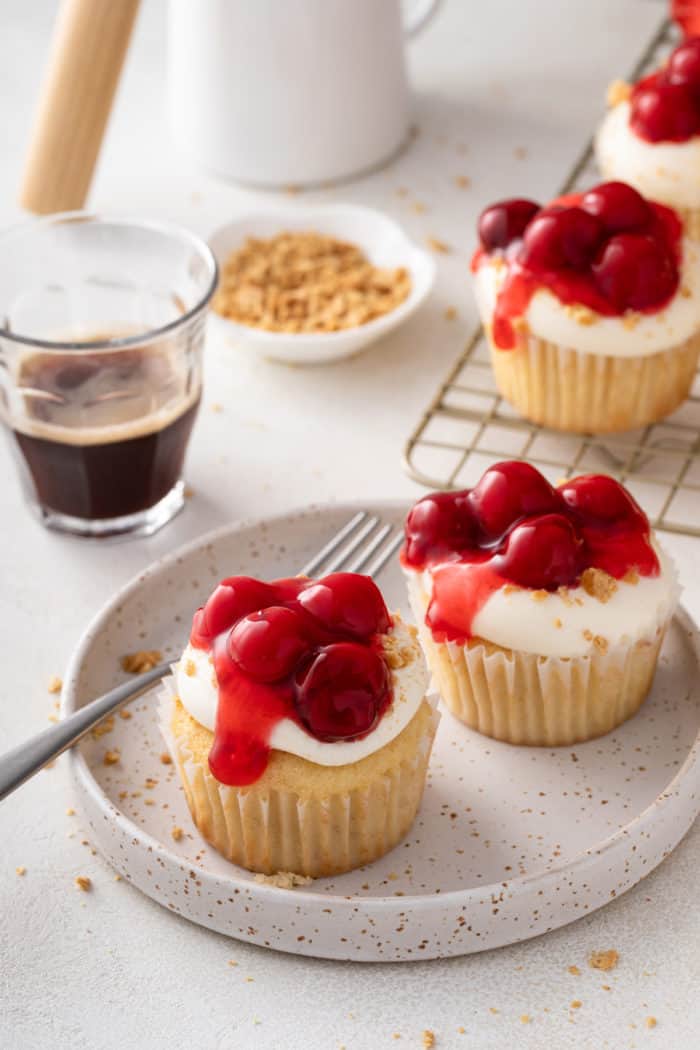 Two cherry cheesecake cupcakes next to a fork on a speckled plate.