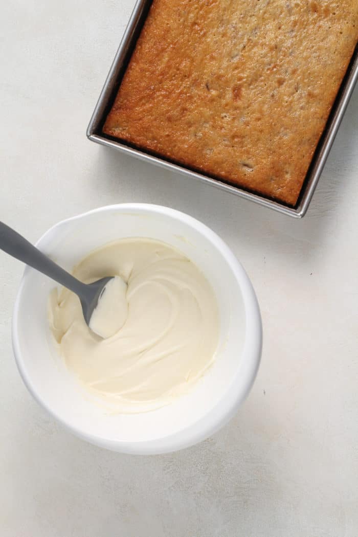 Bowl of cream cheese frosting set next to a baked banana cake.