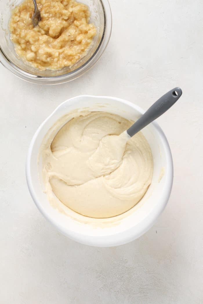 Cake batter in a white mixing bowl next to a bowl of mashed bananas.