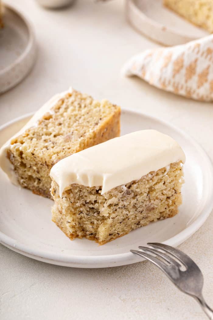 Halved slice of banana cake on a white plate. A bite has been taken from the corner of the cake and a fork is set on the edge of the plate.