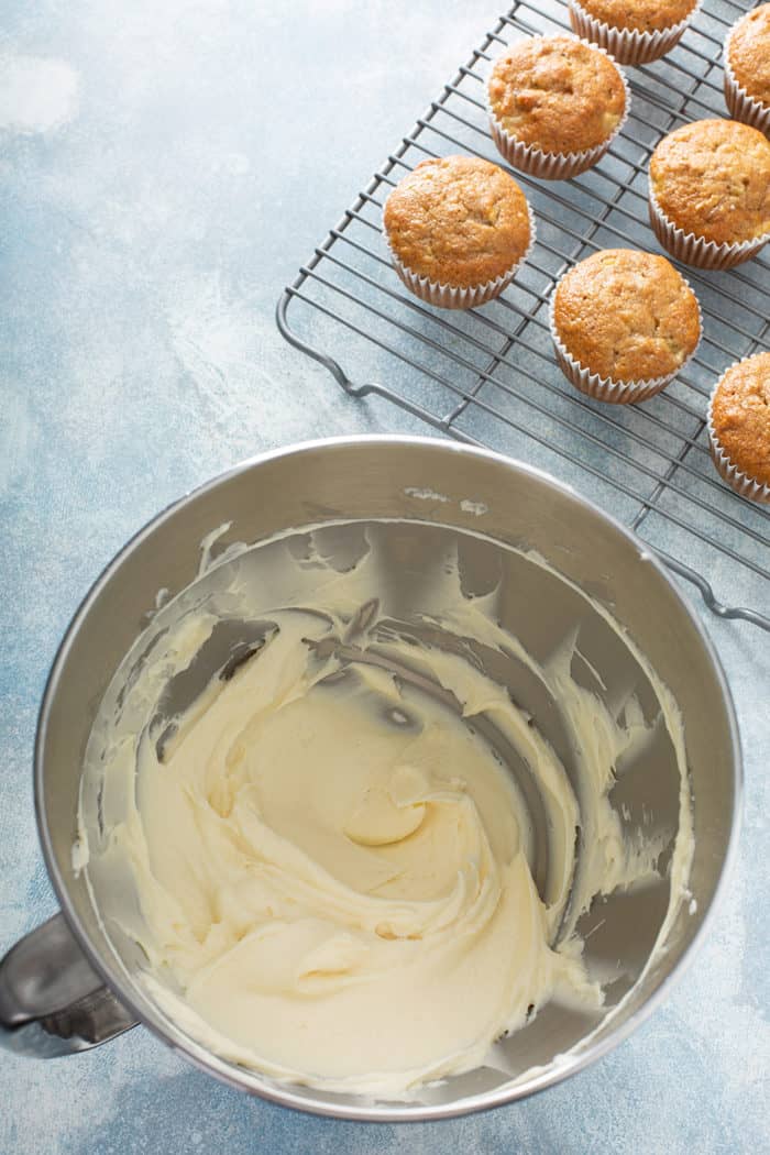 Bowl of cream cheese frosting on a blue countertop next to hummingbird cupcakes on a wire cooling rack