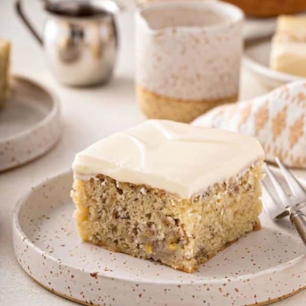 Slice of banana cake topped with cream cheese frosting on a speckled plate.