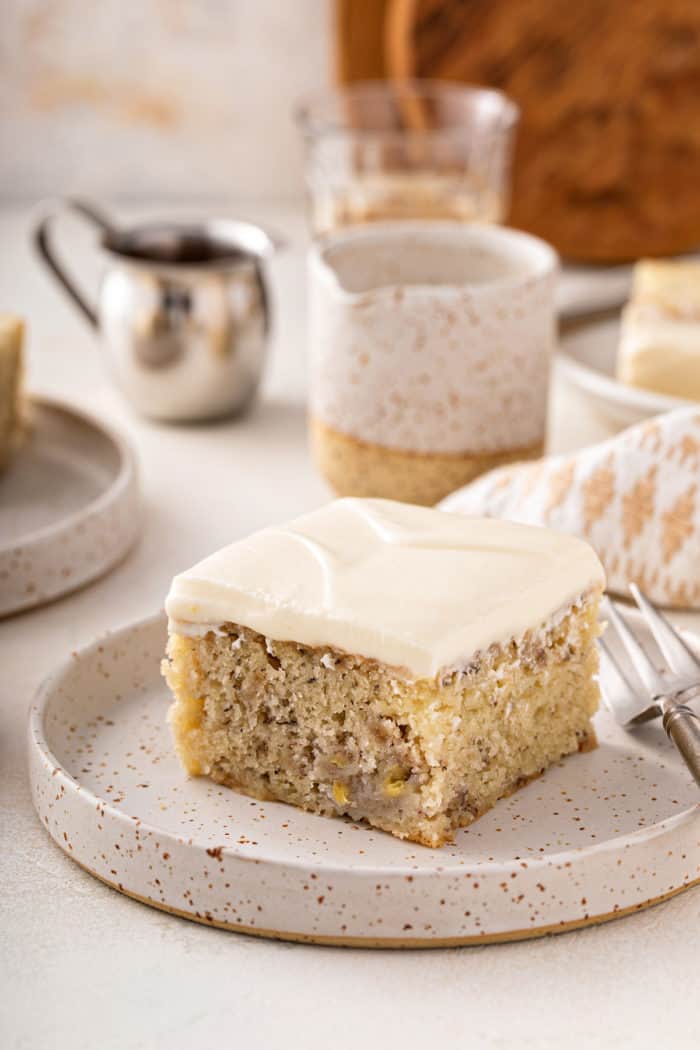 Slice of banana cake topped with cream cheese frosting on a speckled plate.