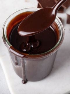 Spoon holding hot fudge sauce over a glass jar of the sauce.