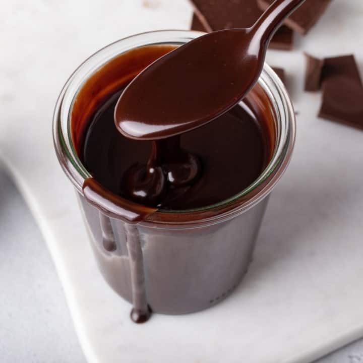 Spoon holding hot fudge sauce over a glass jar of the sauce.