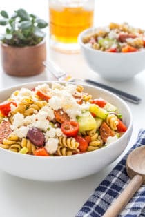 Serving bowl filled with greek pasta salad with a drink and smaller bowl of salad in the background