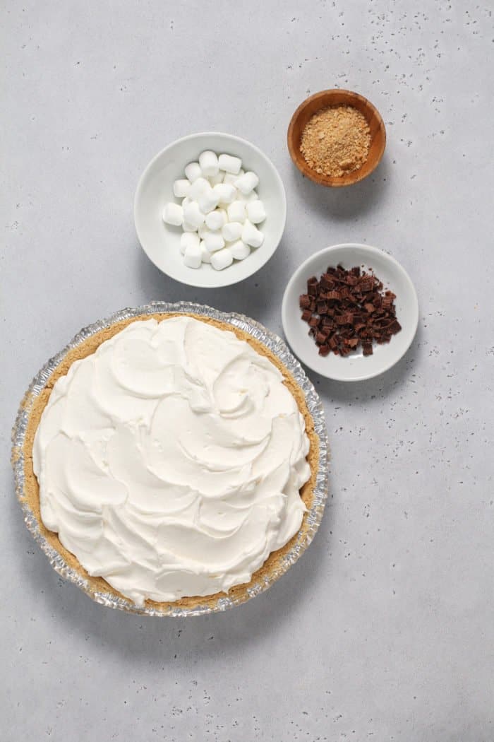 Assembled s'mores pie next to bowls of mini marshmallows, chocolate shavings, and graham cracker crumbs.