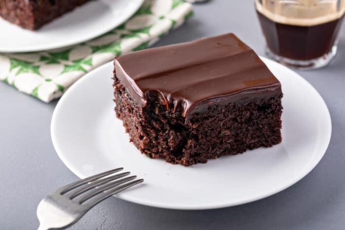 Slice of chocolate zucchini cake set on a white plate. A bite has been taken from the corner of the cake.