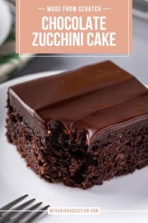 Close up of a slice of chocolate zucchini cake with a bite taken from the corner. A fork is visible perched on the side of the plate. Text overlay includes recipe name.