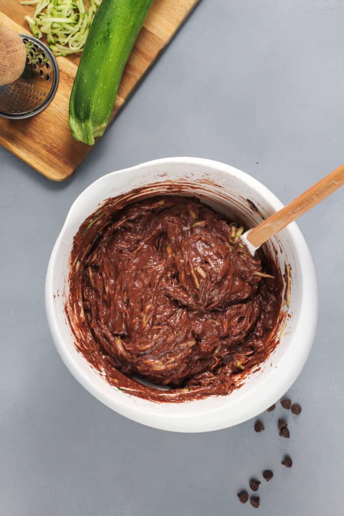 Chocolate zucchini cake batter being stirred in a white mixing bowl.