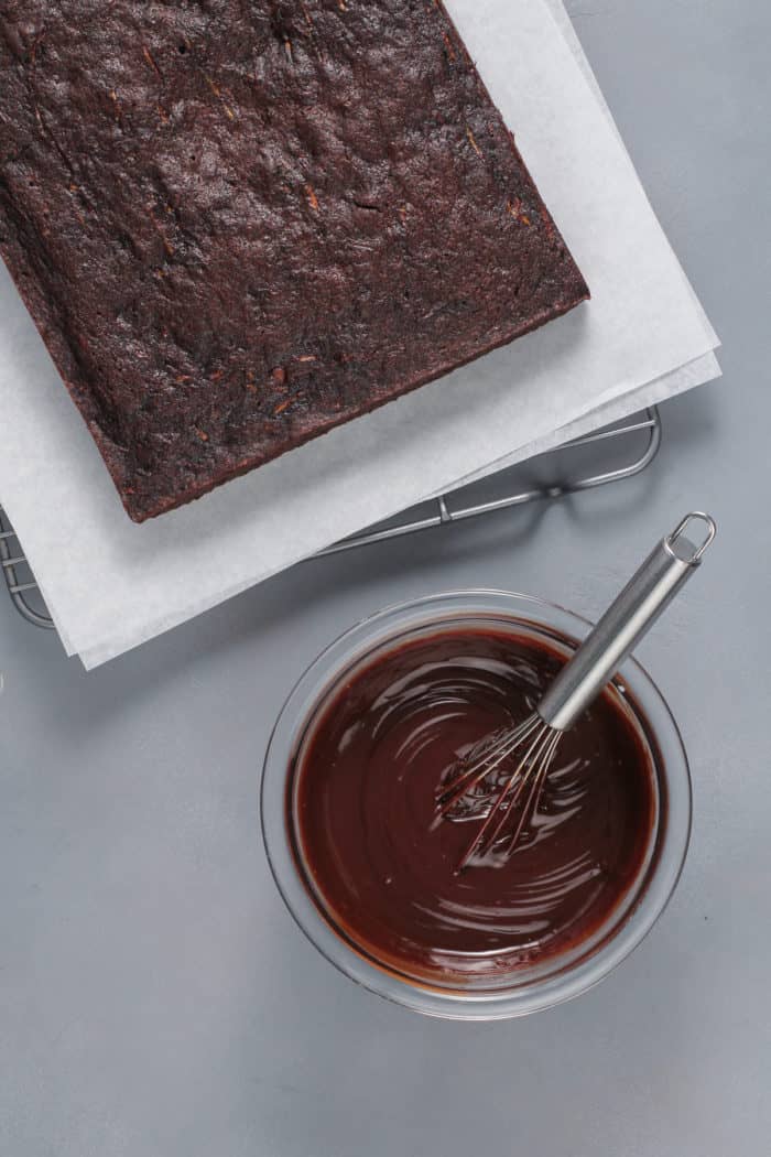 Baked chocolate zucchini cake set next to a bowl of chocolate ganache frosting.