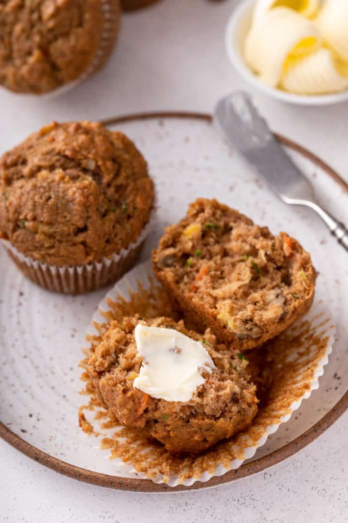 Two morning glory muffins on a plate. One of the muffins is split in half with a pat of butter on one of the halves.