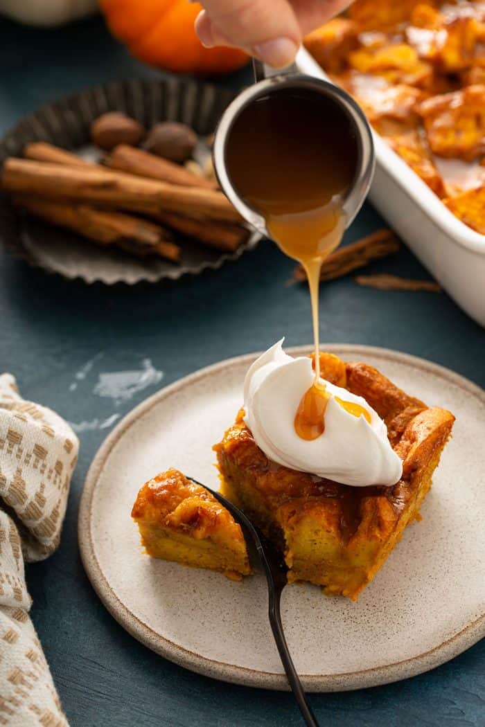 Caramel sauce being drizzled over a slice of pumpkin bread pudding topped with whipped cream