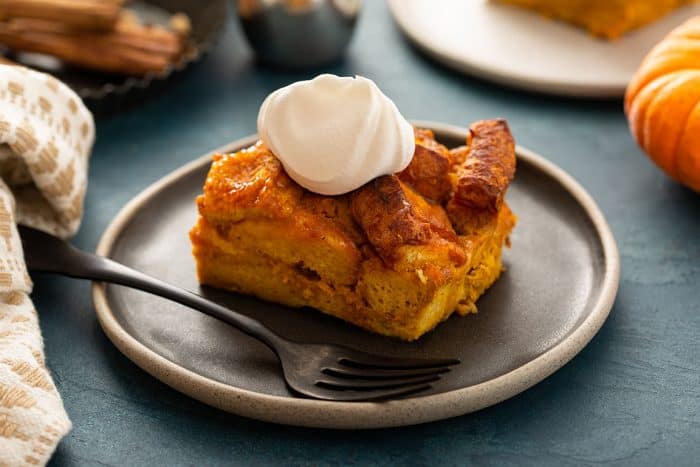 Slice of pumpkin bread pudding topped with whipped cream next to a fork on a plate