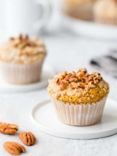 Pumpkin cream cheese muffin on a small round plate next to pecans on a white surface