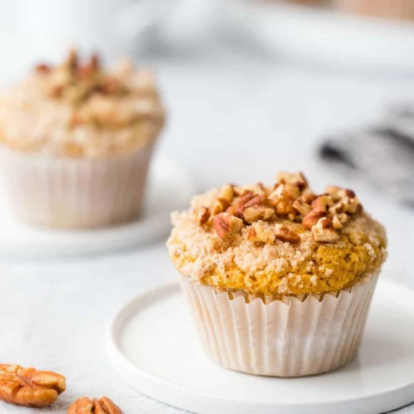 Pumpkin cream cheese muffin on a small round plate next to pecans on a white surface