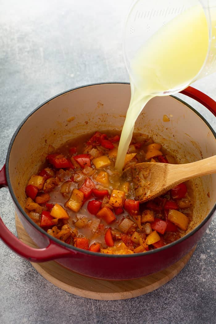 Chicken broth being poured into a dutch oven with sauteed bell peppers and onions