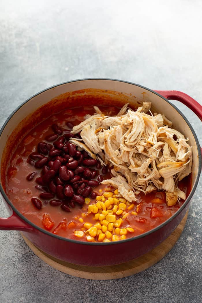 Shredded chicken, beans, and corn being added to a pot of chili in a dutch oven