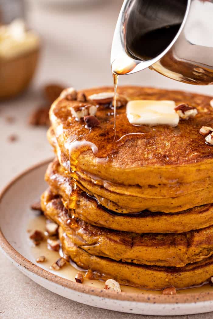 Maple syrup being poured over a stack of pumpkin pancakes on a plate.