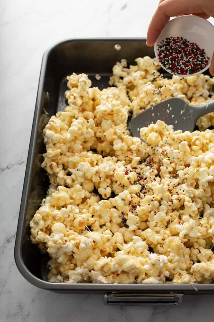 Hand sprinkling holiday sprinkles over sticky popcorn on a baking pan