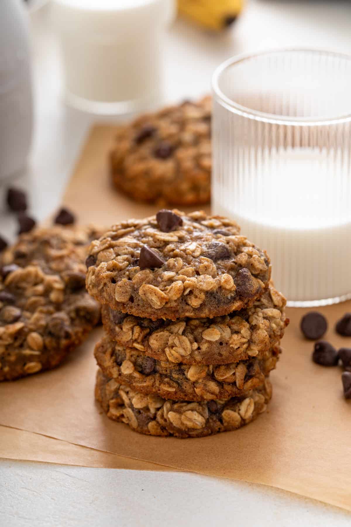 Four banana oatmeal cookies stacked on a piece of parchment paper in front of a glass of milk.
