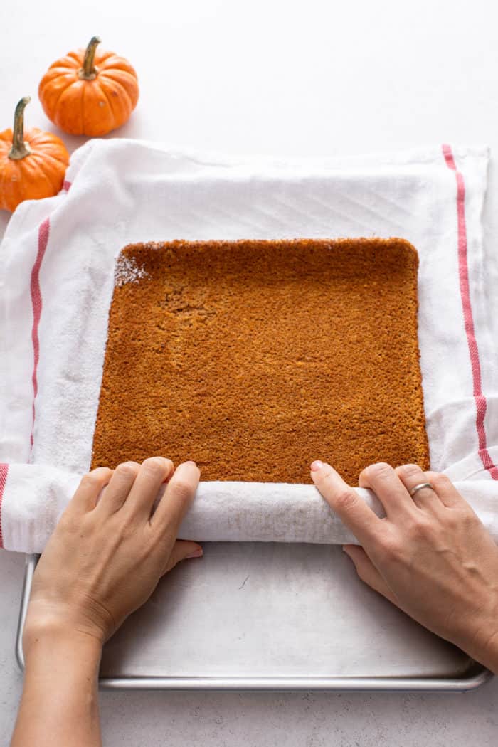 Hands rolling pumpkin sponge cake in a white tea towel.