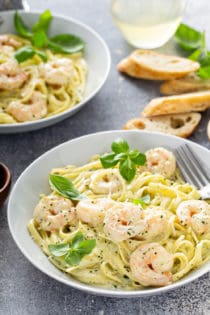 Plated bowl of creamy pesto pasta with shrimp with another bowl and slices of baguette in the background